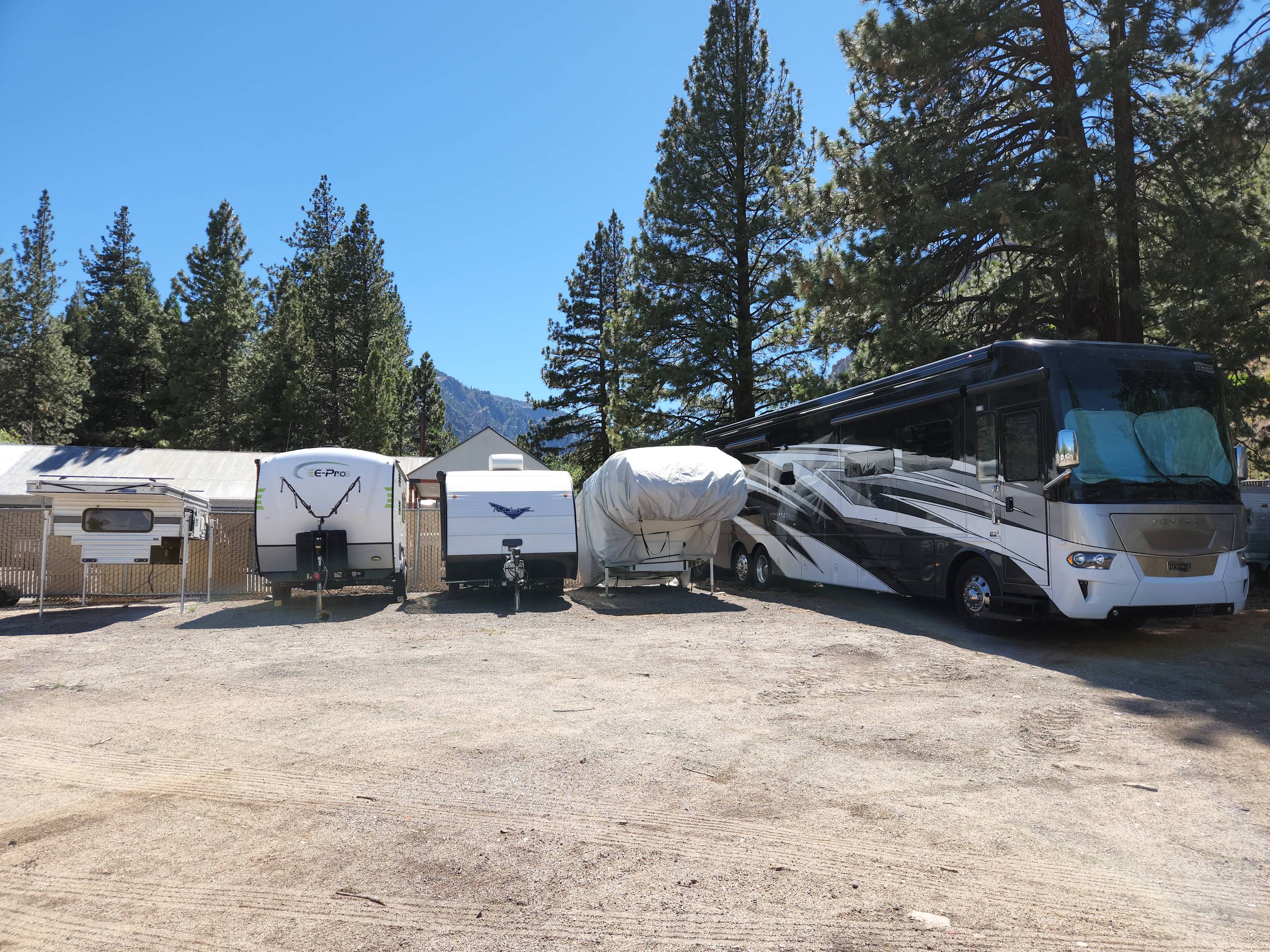 Truck camper, travel trailers, a covered boat and a full-size motorhome parked among pine trees at Tahoe Camper Storage